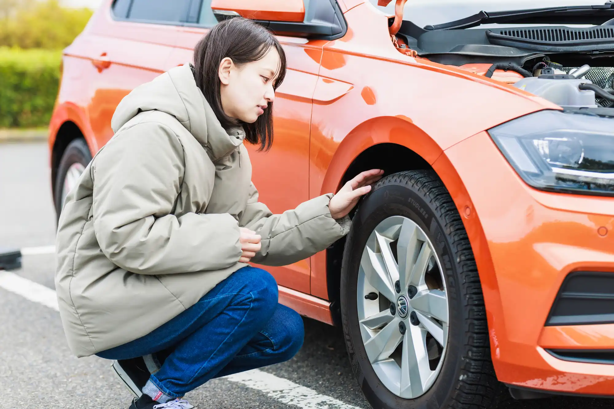 ボンネットを開けたオレンジの車のタイヤを見る女性
