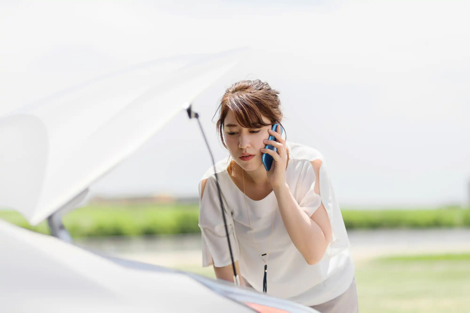 車のボンネットを開けて様子を見ながら電話をする女性